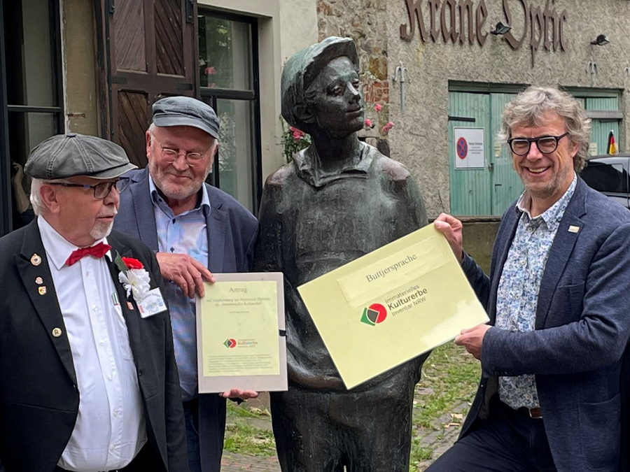 Klaus Siewert, promovierter und habilitierter Sprachwissenschaftler an der Universität Münster (2.v.l.), hat das Logo "Immaterielles Kulturerbe", das 20 Seiten umfassende Antragswerk und den Bewilligungs-Bescheid des NRW-Landesministeriums für Kultur und Wissenschaft an Bürgermeister Michael Jäcke (r.) übergeben. Mit dabei der "Mindener Buttjer", eine Skulptur in der Oberen Altstadt, und Dieter Böhning (l.), der bei Veranstaltungen den Buttjer verkörpert. Foto: Stadt Minden